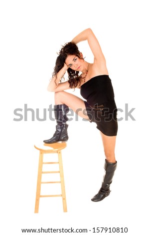 A lovely young slim woman in black shorts and long boots standing for white background with one leg on a chair.