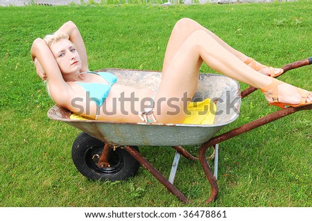 Blond beautiful bikini woman sitting in an old rusty wheelbarrow on a green lawn and enjoying the nice sunshine and relax.
