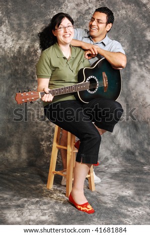 An woman in black pants sitting on a chair with an green guitar for a black  and white background and smiling in the camera, with her husband.