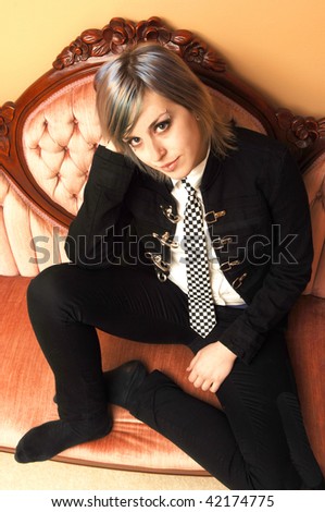 An pretty young girl in a jacket and black and white tie sitting on a pink sofa looking at the camera.