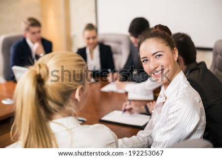 Confident beautiful female team leader sitting in a business meeting at a table with a group of her colleagues turning to smile at the camera, selective focus
