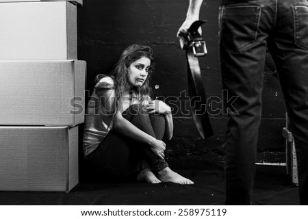 Young woman is a victim of domestic violence and abuse siting on the floor is scared of man with belt on black-and- white background