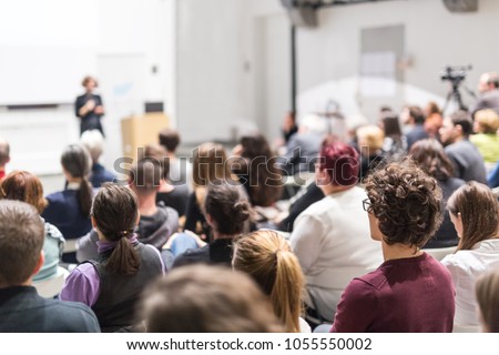 Female speaker giving presentation in lecture hall at university workshop. Audience in conference hall. Rear view of unrecognized participant in audience. Scientific conference event.