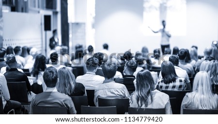 Speaker giving a talk in conference hall at business event. Focus on unrecognizable people in audience. Business and Entrepreneurship concept. Blue toned greyscale image.