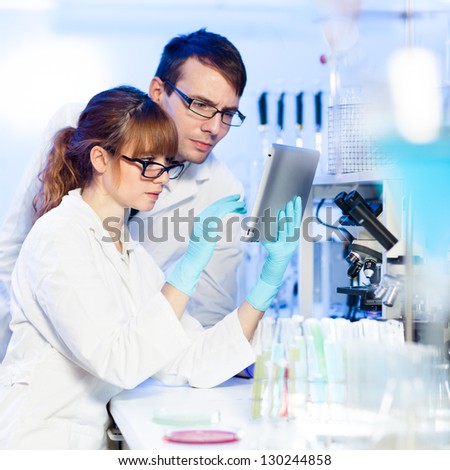Attractive young research scientist and her post doctoral male supervisor looking focused at the tablet in the life science (forensics, microbiology, biochemistry, genetics, oncology...) laboratory.