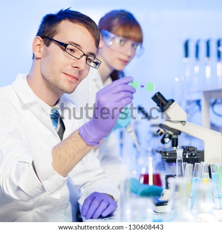 Young male researcher looking at the microscope slide in the life science (forensics, microbiology, biochemistry, genetics, oncology ) laboratory. Female assistant scientist working in the background.