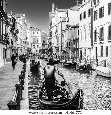 Gondolas Passing On Small Canal Among Old Historic Houses And Bridge In Venice, Italy. Black And White Image.
