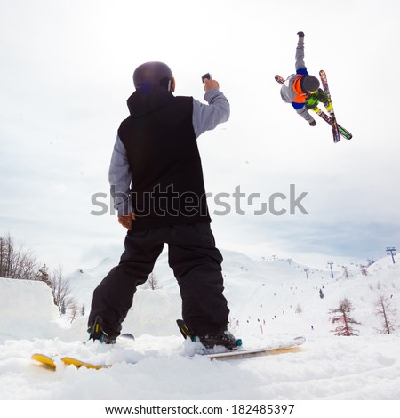 Free style skier performing a high jump. Buddy filming it on a Gopro camera. Ski lifts in the mountains in the background.