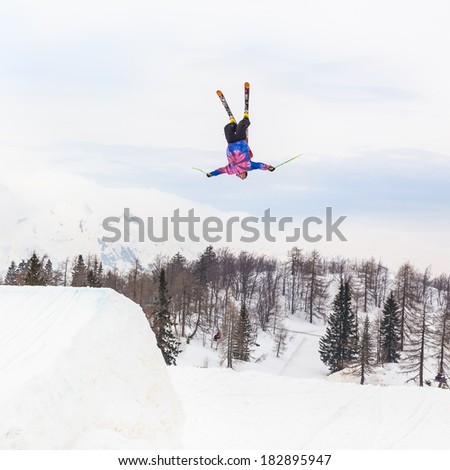 Free style skier performing a high back flip jump. Ski lifts in the mountains in the background.