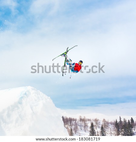 Free style skier performing a high up in the sky back flip jump. Snow covered mountains in the background.