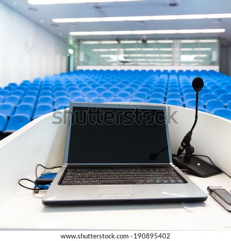 Laptop and microphone on the rostrum in empty conference hall with blue velvet chairs.