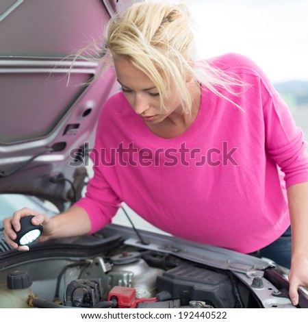 Self-sufficient confident modern young woman inspecting broken car engine with a flashlight in her hand.