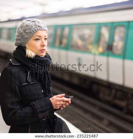 Young woman in winter coat with a cell phone in her hand waiting on the platform of a railway station for train to arrive. Public transport.