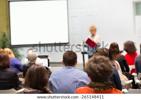 Female speaker giving presentation in lecture hall at university. Participants listening to lecture and making notes.