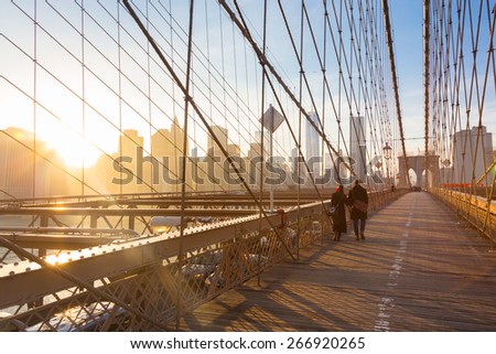 Couple walking on pedestrian path across Brooklyn bridge. New York City Manhattan downtown skyline in sunset with skyscrapers illuminated over East River panorama as seen from Brooklyn bridge.