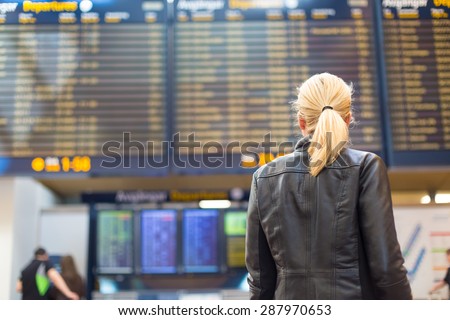 Casually dressed young stylish female traveller checking a departures board at the airport terminal hall in front of check in couters. Flight schedule display blured in the background. Focus on woman.