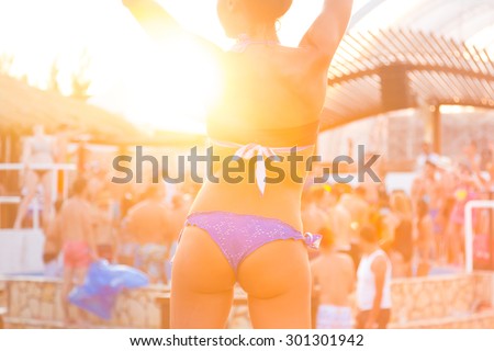 Sexy hot girl wearing brazilian bikini dancing on a beach party event in sunset. Crowd dancing and partying at poolside in background. Summer electronic music festival. Hot summer party vibe.