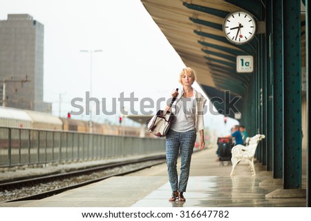 Blonde caucasian woman waiting at platform of railway station bearing vintage shoulder bag.