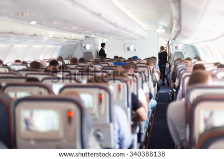 Interior of large passengers airplane with people on seats and stewardess in uniform walking the aisle.
