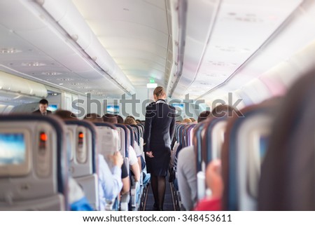 Interior of airplane with passengers on seats during flight. Stewardess in dark blue uniform walking the aisle. Horizontal composition.