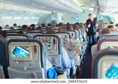 Interior of airplane with passengers on seats during flight. Steward in dark blue uniform walking the aisle. Horizontal composition.