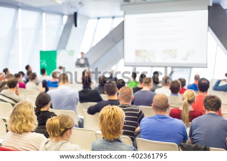 Man giving presentation in lecture hall. Male speeker having talk at public event. Participants listening to lecture. Rear view, focus on people in audience.