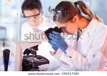 Life scientists researching in laboratory. Female young scientist and her post doctoral supervisor microscoping in their working environment. Health care and biotechnology.