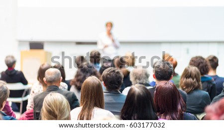 Business and entrepreneurship symposium. Female speaker giving a talk at business meeting. Audience in conference hall. Rear view of unrecognized participant in audience. Copy space on white screen.