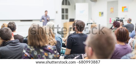 Male speaker giving presentation in lecture hall at university workshop. Audience in conference hall. Rear view of unrecognized participant in audience. Scientific conference event.