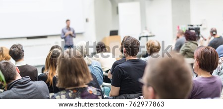 Male speaker giving presentation in lecture hall at university workshop. Audience in conference hall. Rear view of unrecognized participant in audience. Scientific conference event.