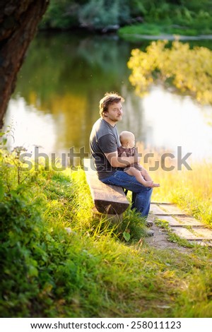 Middle age father with his little baby sitting on wooden bench at the sunset