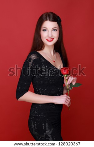 Charming young woman in a black dress holding a single red rose on a red background