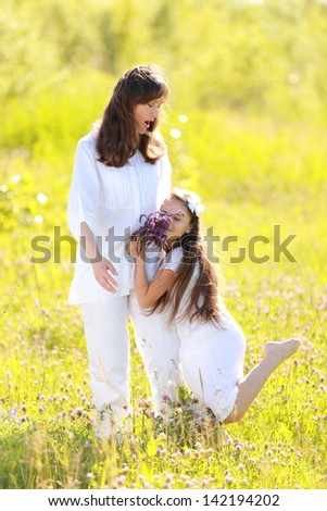 Smiling and cute mother and daughter in summer clothes holding wildflowers walking and laughing in the park outdoors