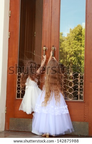 Two charming little girls in long white dresses stand near the mirrored doors of the building outdoors