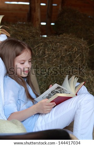 Beautiful little girl with long hair reading a book in the hayloft on a horse farm