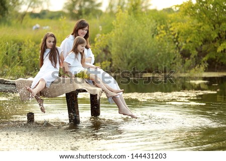 Cheerful Young Woman With Two Young Girls Sitting On A Pier At The Lake On The Background Of Beautiful Nature In Spring