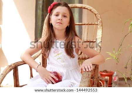 Cute little girl in a white dress sits in a wicker rocking chair and holding a red apple outdoors