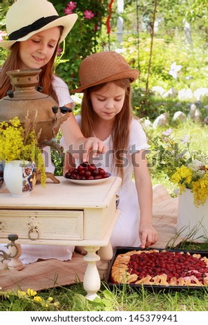 Two smiling young girls in retro hats drinking tea from a Russian samovar in the summer garden and eat a cherry/Russian national traditions