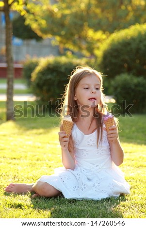Contented smiling little girl is holding two ice creams sitting on the grass on a hot summer day