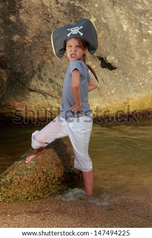 Pretty little girl in a hat with a skeleton pirate symbol of piracy is in the water near a huge rock by the sea