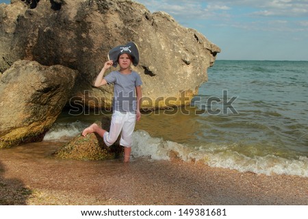 Beautiful little girl in a hat with a skeleton pirate symbol of piracy is in the water near a huge rock by the sea
