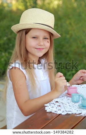 Cute little girl in a summer hat playing with children\'s dishes in the back yard of a house outdoors