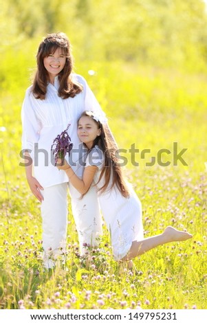 Smiling and cute mother and daughter in summer clothes holding wildflowers walking and laughing in the park outdoors