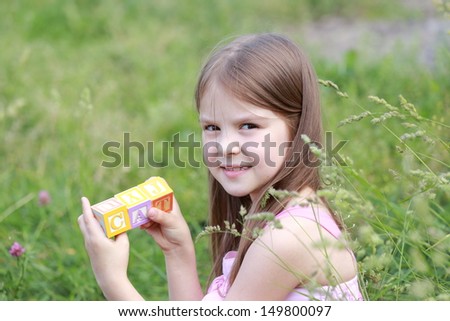 Young girl with long hair sitting in spring grass playing with blocks with letters and form words outdoors