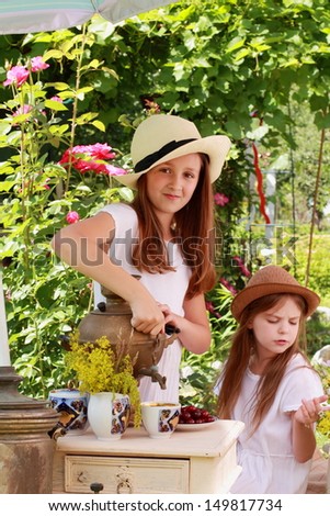 Adorable little girl drinking tea from an antique Russian samovar in the summer garden in the street / Russian antiques and national traditions