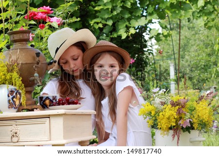 smiling young girls in retro hats drinking tea from a Russian samovar in the summer garden and eat a cherry/Russian national traditions