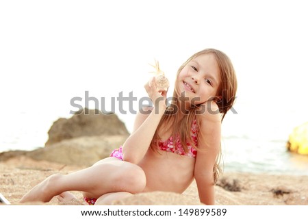 Cute smiling little girl in a bathing suit sits on a background of sea sand with a beautiful sea shell on the beach