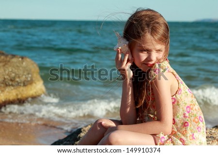 Portrait of a happy young girl in a summer dress with a seashell sits on the shore of the Black Sea on a hot sunny day