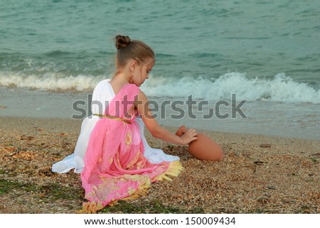Smiling cheerful little girl in a pink toga holding an antique amphora and is barefoot on the beach/Girl in the role of the Roman goddess outdoors