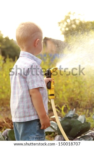 Cute little blond boy in summer clothes watering the lawn with a hose in a hot summer day